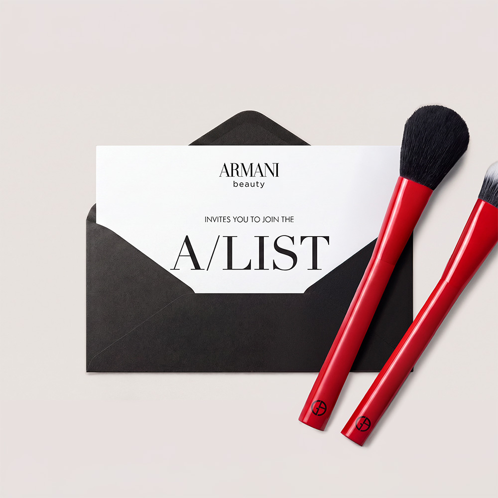 Armani Beauty A-List invitation beside two red makeup brushes.